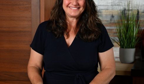 Smiling woman with dark hair in a black top holding papers, standing at a marble table in a room with wooden and plant decor.