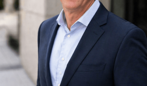 Man in a dark suit and light blue shirt standing outdoors, smiling, with a building in the background.