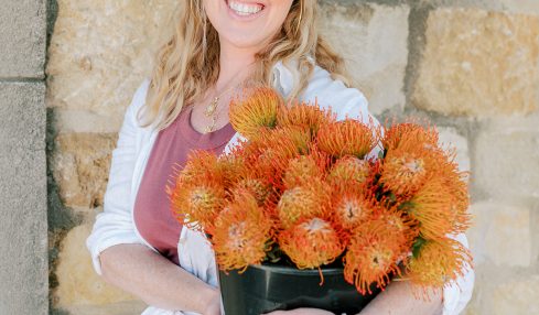 Woman with blonde hair holding a black pot of orange flowers, standing against a stone wall.