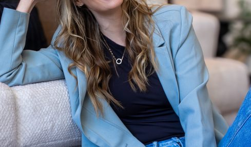 Young woman with long wavy hair smiling, sitting on a sofa in a casual setting, wearing a blue blazer and jeans.